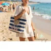 Una mujer sonriente en la playa lleva colgada al hombro la bolsa de rayas marineras para profesora con el texto "Nuestra PROFE Marisa tiene mucha CLASE". Viste un vestido playero claro, gafas de sol y lleva un libro en la mano, con el mar y la arena de fondo.