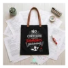 Bolsa de tela negra para profesora con frase divertida sobre corregir examenes y asas marrones, estirada sobre mesa de examenes